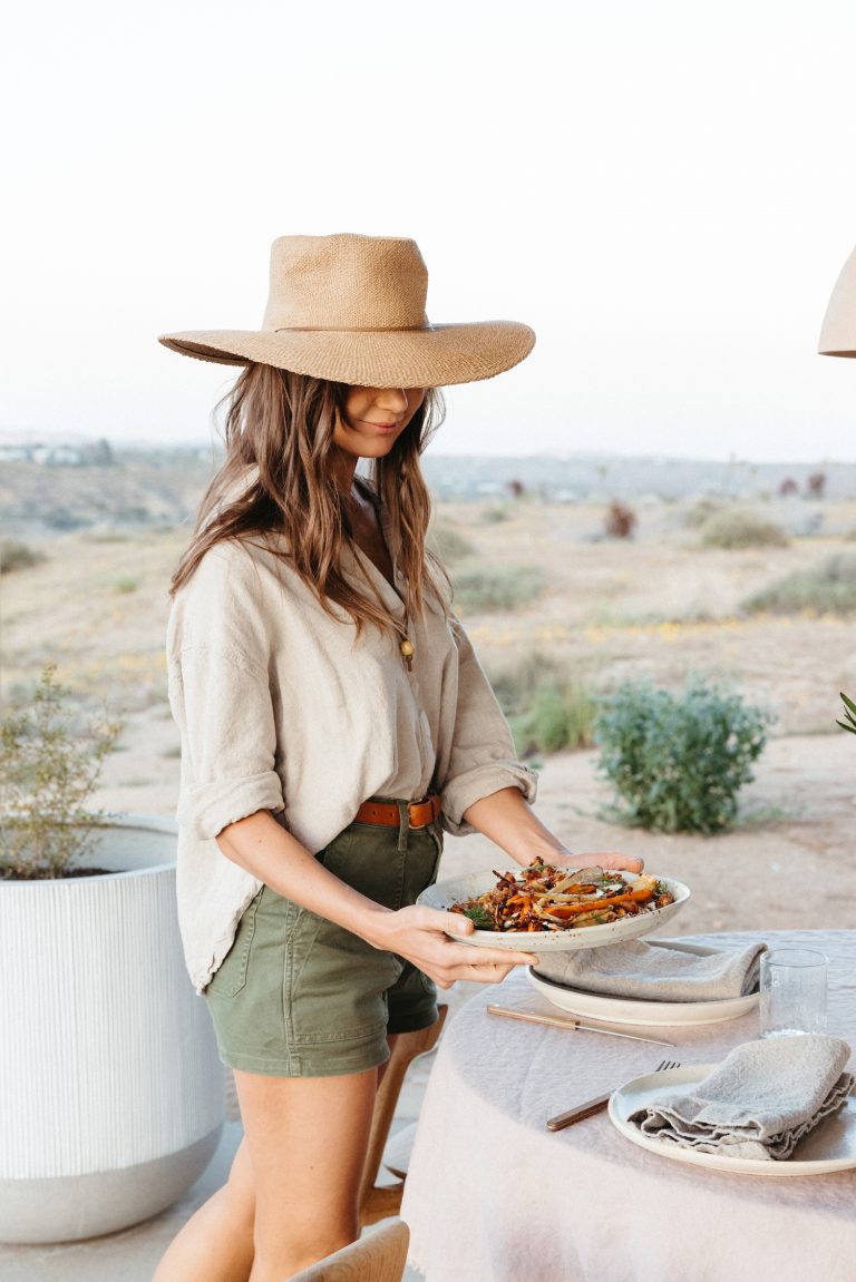 Brunette woman wearing sun hat serving a plate of roasted carrots outdoors.